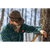 Photo of man in the woods tapping a maple tree for syruping. Man is wearing a brown Stormy Kromer Waxed Cotton Cap and a blue and green plaid flannel overshirt.