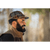 Image of bearded man in his 30s with trees blurred out in the background. Man is wearing a plaid Stormy Kromer Rancher Cap and a vest.
