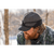 Image of bearded man in his 40s in front of a stand of trees in winter and looking down. Man is wearing a Stormy Kromer Rancher cap in charcoal and a plaid jacket.