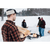 Back view of a man carrying logs to an outdoor fire pit in the winter. The man is wearing a dark Stormy Kromer Bergland cap and a rust and navy plaid shirt. 3 figures stand around the fire in the background.