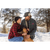 Photo of a man and woman kneeling down with their arms around their dog on a snowy path with trees in the background. The man is wearing a dark Stormy Kromer Bergland Cap and a green plaid overshirt. The woman is wearing a black Stormy Kromer cap and a red/black plaid jacket.