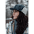 Close up photo of a woman with long dark hair wearing a gray plaid Snowdrift Cap with the earflaps tied up on top. There are blurry trees in the background and she is wearing a gray plaid jacket.