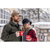Photo of man and woman standing with arms around each other, holding coffee mugs in front of a large porch with stone railing and lots of windows in the winter. Man is wearing Stormy Kromer Insulated Waxed Cotton Cap in tan with olive wool coat. Woman is wearing red and black check jacket and black cloche style hat.