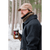 Image of a man standing in front of a very wooded area with snow on the ground holding a cup of coffee and looking to the side. Man is wearing a Stormy Kromer Marsh Cap with the earflaps folded down to cover his ears and a dark gray wool coat.