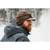 Side profile view of a man with a red beard wearing a dark brown waxed Rancher Cap with the earband pulled down over his ears. It is a very snowy scene with trees and blowing snow.