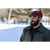 Bearded man in his 30s looking off in the distance with snow and trees behind him. Man is wearing a red and black plaid Stormy Kromer Original Cap with Leather and wool coat.