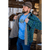 Photo of man putting on his green plaid Camp Shirt Jack over a bright blue shirt with a trout image. He is wearing a brown Stormy Kromer cap and is standing in front of a wood pile.