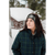 Woman with long dark hair standing outside in the snow in front of log cabin wearing a green and blue plaid Stormy Kromer jacket and a gray and white Stormy Kromer headband.