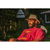 Photo of a bearded man relaxing in a lawn chair in the sun wearing a red shirt with a deer graphic and wearing The Mesh Hiker hat in Khaki. There is a house and an old beer sign in the background.