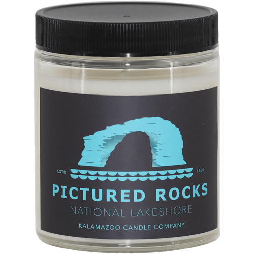 Glass jar candle from Kalamazoo Candle with black lid and label, printed with a Pictured Rocks drawing in bright blue
